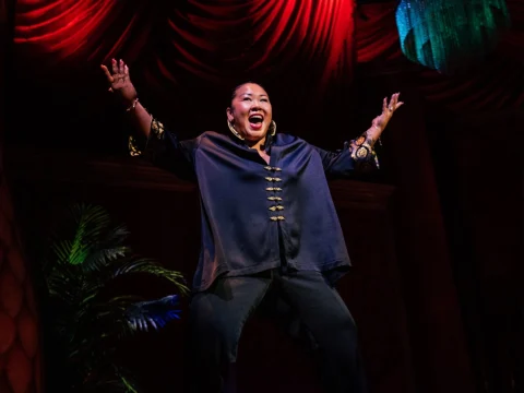 Person on stage with arms raised, smiling widely under dramatic red lighting, wearing a dark shirt with decorative buttons.