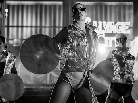 A performer in futuristic attire and sunglasses holds large discs on stage, with "SALVAGE CITY" illuminated in the background.