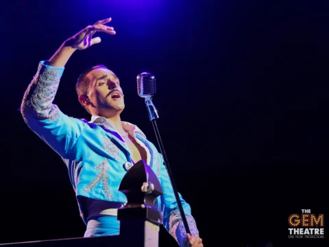 A performer wearing a blue suit sings into an old-fashioned microphone on stage.