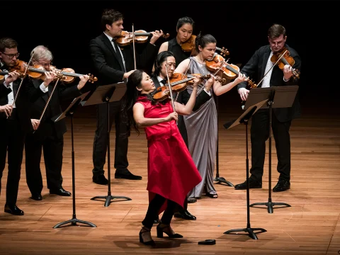 A group of musicians performing on stage with violins. The central violinist in a red dress is playing passionately, with other musicians in the background dressed in darker colors. Music stands are visible.