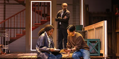 Three people are on a stage set resembling a porch; two sit and examine papers while a third stands in the background with arms crossed, watching them.