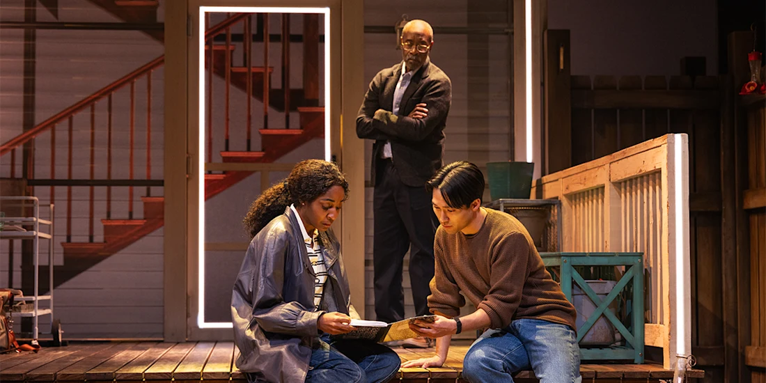 Three people are on a stage set resembling a porch; two sit and examine papers while a third stands in the background with arms crossed, watching them.