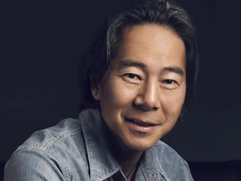 A man with straight, dark hair wears a denim shirt and smiles slightly, facing the camera against a dark background.