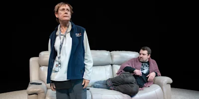 An older person stands in front of a couch while a younger person sits on the couch, wrapped in a blanket, against a black background.