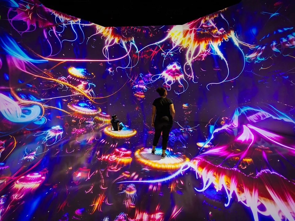 Two people stand on illuminated platforms in a vibrant, immersive digital art exhibit with swirling neon colors and abstract shapes.