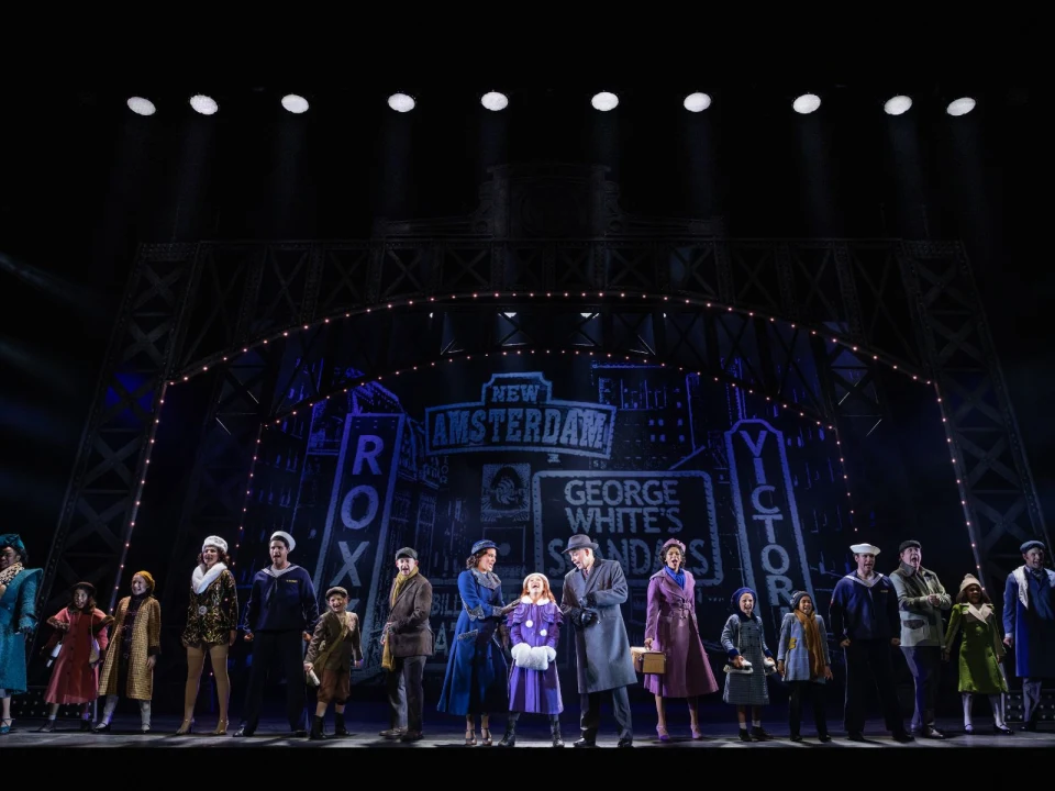 Cast members stand on stage during a theater production, in front of a backdrop featuring vintage Broadway signs from "New Amsterdam," "Roxy," "George White's," and "Victoria.