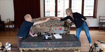A man kneels on the floor reaching toward a woman standing beside a couch in a living room with patterned furniture and wooden floors.