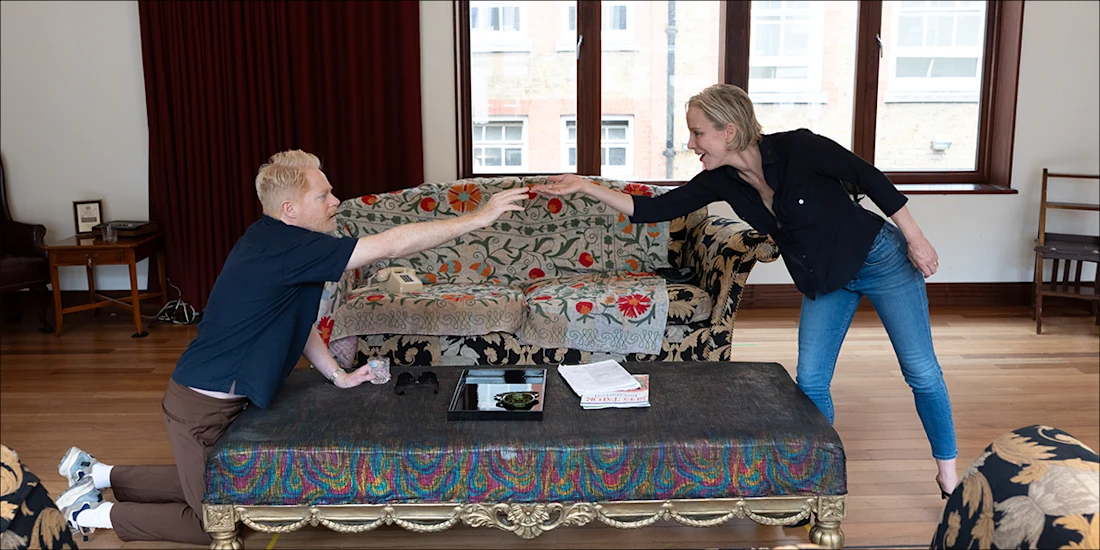 A man kneels on the floor reaching toward a woman standing beside a couch in a living room with patterned furniture and wooden floors.