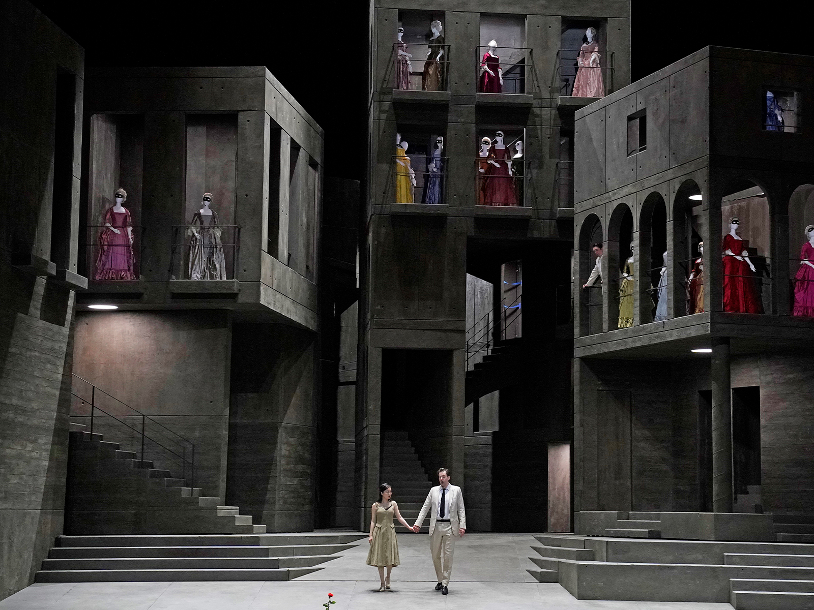 Two people hold hands on stage in front of a gray, multi-level set, while numerous individuals in period costumes stand on balconies and in windows above.