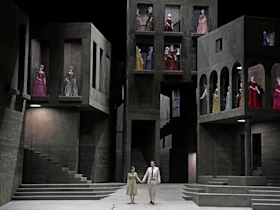 Two people hold hands on stage in front of a gray, multi-level set, while numerous individuals in period costumes stand on balconies and in windows above.