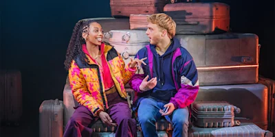 Two people in colorful jackets sit on stacked suitcases, engaged in conversation, with one holding a phone and gesturing animatedly.