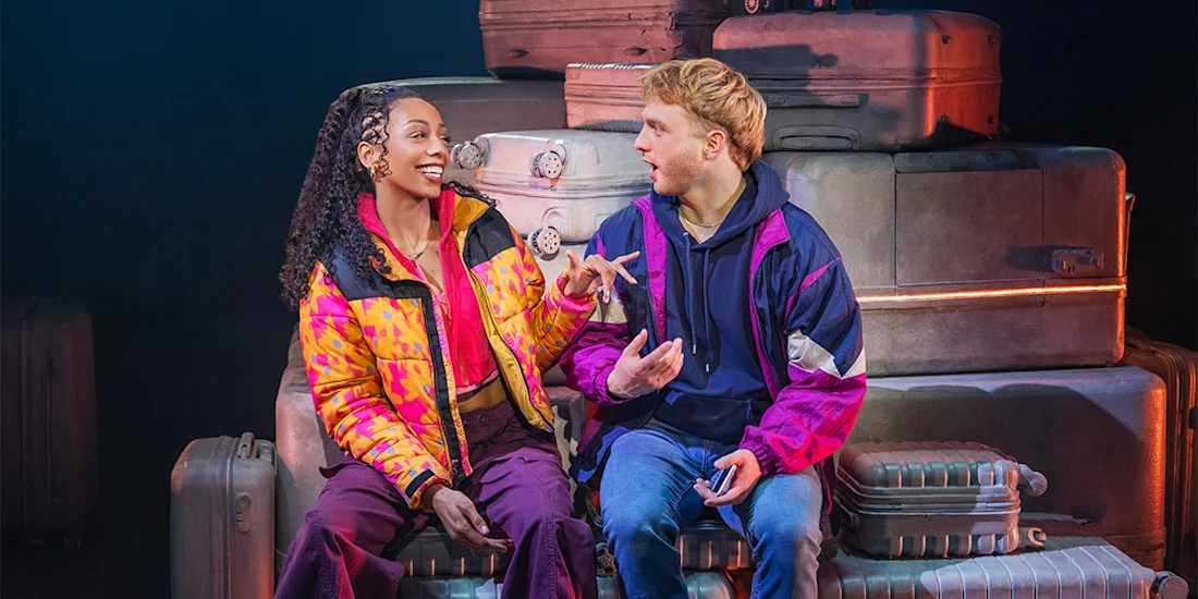 Two people in colorful jackets sit on stacked suitcases, engaged in conversation, with one holding a phone and gesturing animatedly.