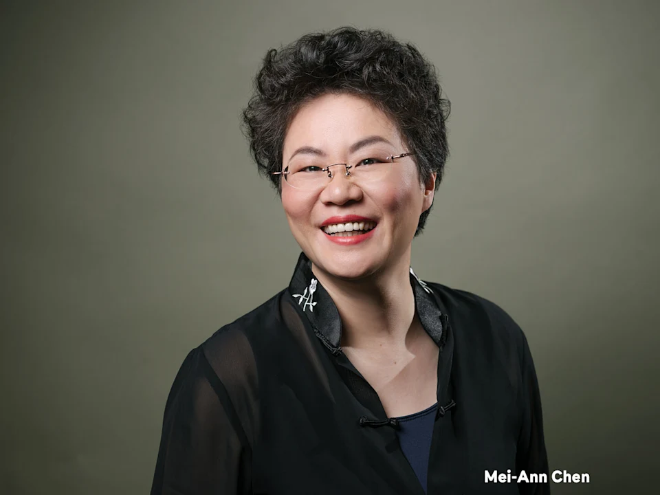 A woman with short curly hair, glasses, and a black top smiles in front of a plain background. The name Mei-Ann Chen appears in white text at the bottom right.