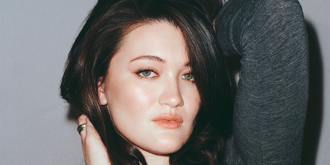 A person with long brown hair and green eyes poses with one hand behind their head, wearing a dark long-sleeve top and a ring on one finger, against a plain background.