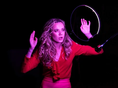 A woman in a red outfit stands with raised hands in front of a lit ring light, illuminated by purple lighting against a dark background.