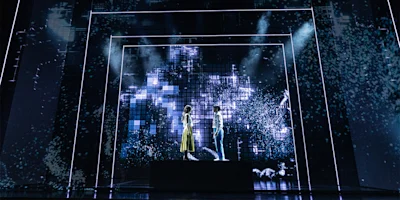 Two people stand facing each other on a platform, surrounded by blue digital light effects and geometric patterns on a dark stage.