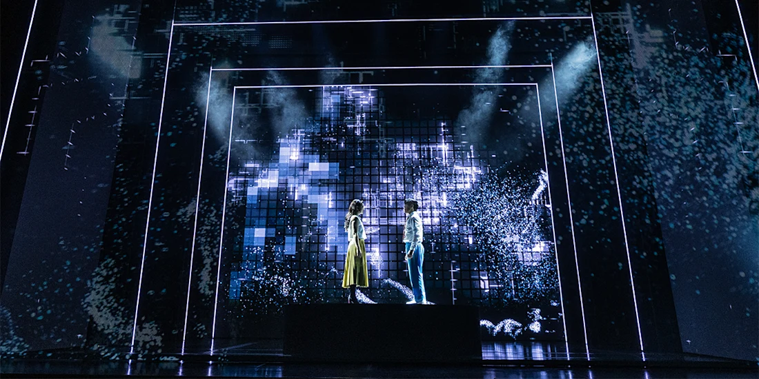 Two people stand facing each other on a platform, surrounded by blue digital light effects and geometric patterns on a dark stage.