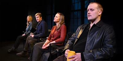 Four people sit in a row of chairs on stage, facing forward. Three look attentive, while one holds a yellow mug. Large windows are visible in the background.