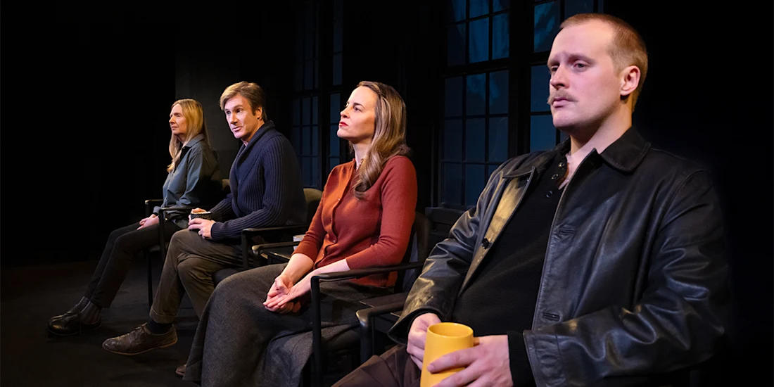 Four people sit in a row of chairs on stage, facing forward. Three look attentive, while one holds a yellow mug. Large windows are visible in the background.