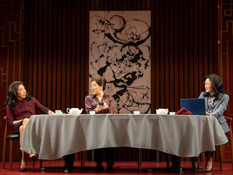 Three women sit at a table covered with a gray tablecloth, having a conversation. Cups, a teapot, and a laptop are on the table. An abstract artwork hangs on the wall behind them.