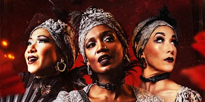Three women in ornate, vintage-inspired costumes and headpieces pose dramatically against a red background.