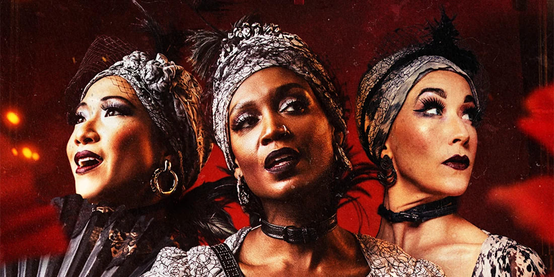 Three women in ornate, vintage-inspired costumes and headpieces pose dramatically against a red background.