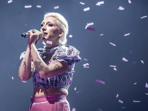 A singer with short blond hair performs on stage, holding a microphone, surrounded by falling confetti under purple lighting.