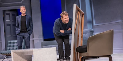 Two men are on stage; one stands in the background while the other closely inspects a large framed object leaning against a chair.