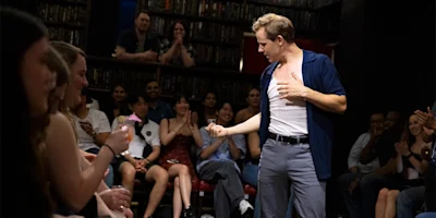 A man performs in front of an audience seated on both sides, holding a small glass and gesturing with his other hand, in a room lined with bookshelves.
