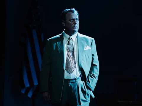A man in a suit stands with one hand in his pocket under dramatic lighting, with an American flag visible in the background.