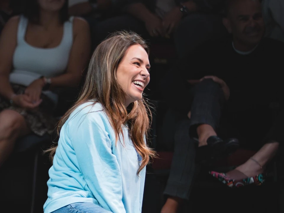 A woman in a light blue sweater is sitting and smiling, surrounded by people in a dimly lit setting.