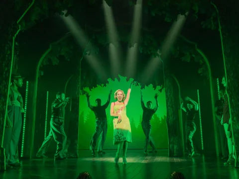 A woman in a green dress sings on stage, surrounded by dancers, under green lighting with a forest backdrop.