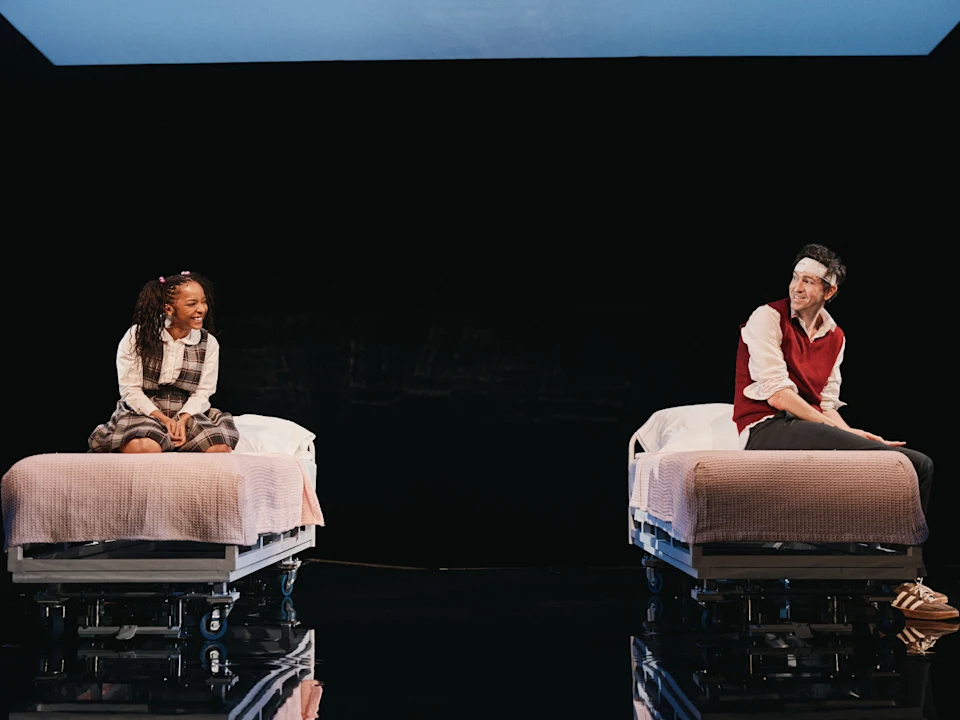Production photo of Gruesome Playground Injuries in New York, showing a young girl and an adult sit on separate hospital beds on a dark stage, facing each other and smiling.