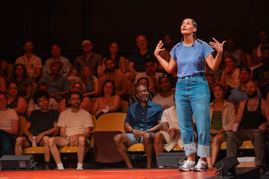 A person in a blue shirt and jeans performs on stage, gesturing with raised hands, while a seated audience watches attentively in the background.