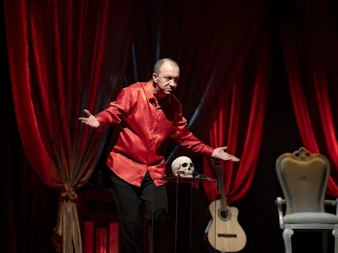 A man in a red shirt stands on stage with arms outstretched next to a human skull on a small table; a guitar and chair are in the background.