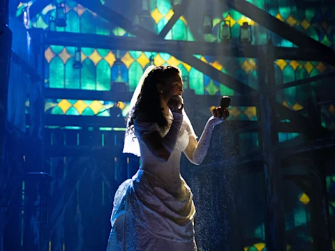 A woman in a white wedding dress and veil stands on a stage, holding an apple in one hand and a small mirror in the other, with a stained-glass backdrop.