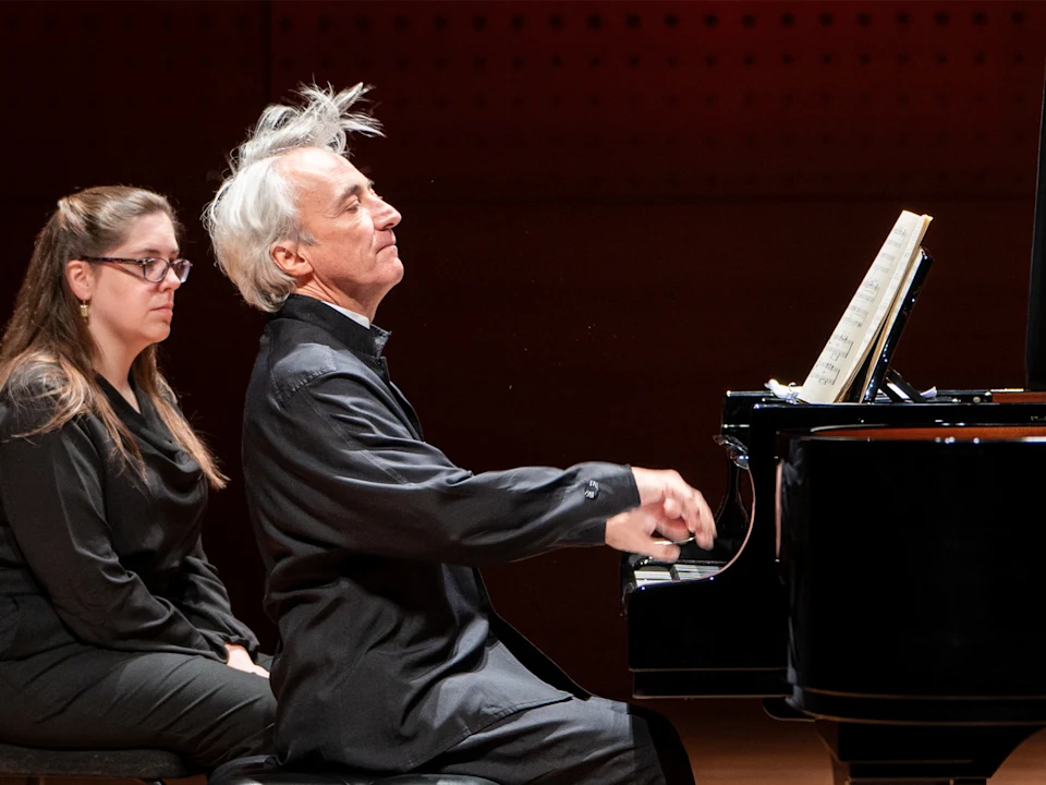 Photo of Jean-Efflam Bavouzet playing a grand piano with sheet music in front of him while a woman sits nearby, watching attentively.