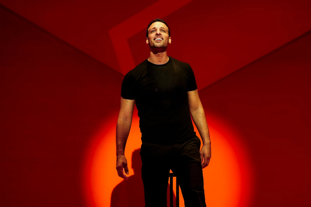 Production photo of Other in New York, showing a man in a black t-shirt and pants stands on stage under a spotlight, with a red background and geometric shapes behind him.