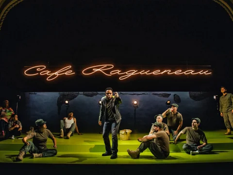 A theater performance shows several actors on stage, some seated and some standing, with a neon sign reading "Cafe Ragueneau" in the background.