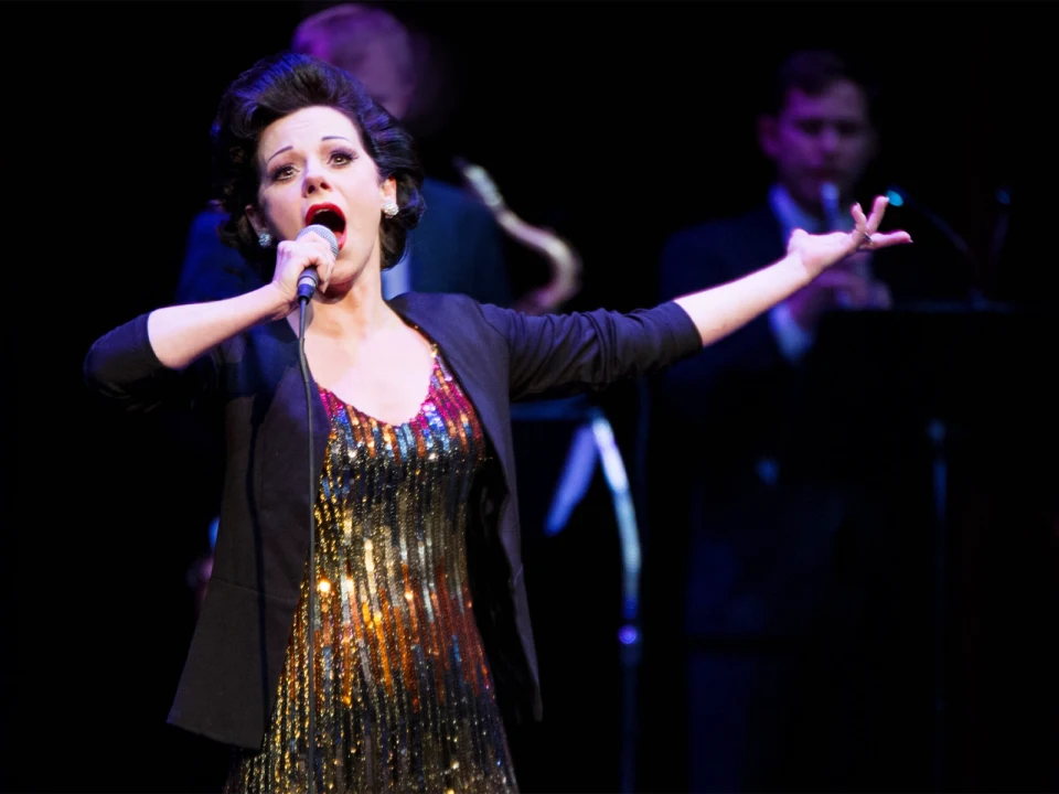 Woman in a glittery dress and black blazer sings passionately on stage, holding a microphone with an orchestra blurred in the background.