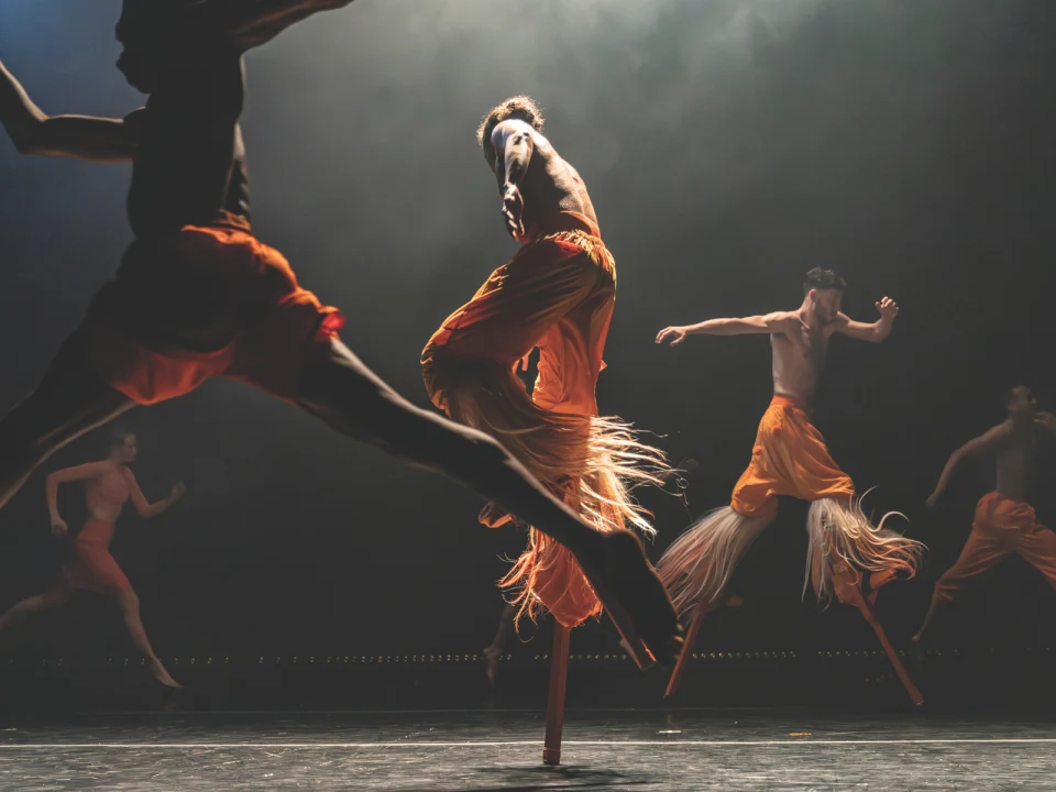 Three dancers in bright orange costumes perform dynamic mid-air movements on stage against a dark backdrop.