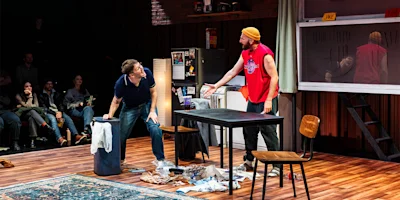 Two men engage in a heated conversation in a cluttered apartment set on stage, with an audience watching in the background.