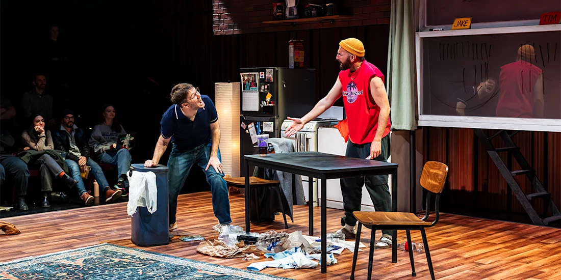 Two men engage in a heated conversation in a cluttered apartment set on stage, with an audience watching in the background.