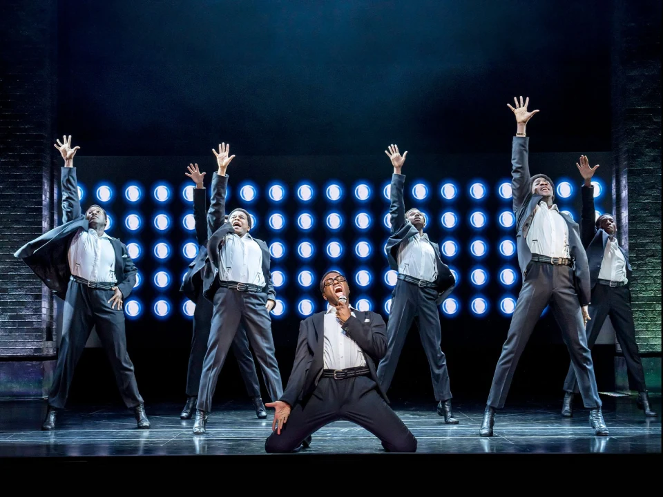 Six performers in suits strike dynamic poses on stage during a musical number, with bright circular lights in the background.