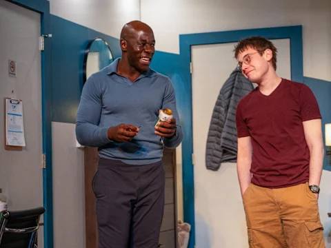 Two men standing in a room; one is holding a jar and smiling, while the other is wearing glasses and smirking. Coats hang on the wall behind them.