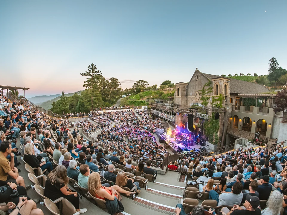 A large audience watches a live outdoor concert at a hillside amphitheater during the evening, with a scenic backdrop of trees and a clear sky.