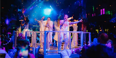 Performers dressed in white sing and dance on a stage with bright lights, confetti, and an audience watching in a festive, nightclub-like setting.