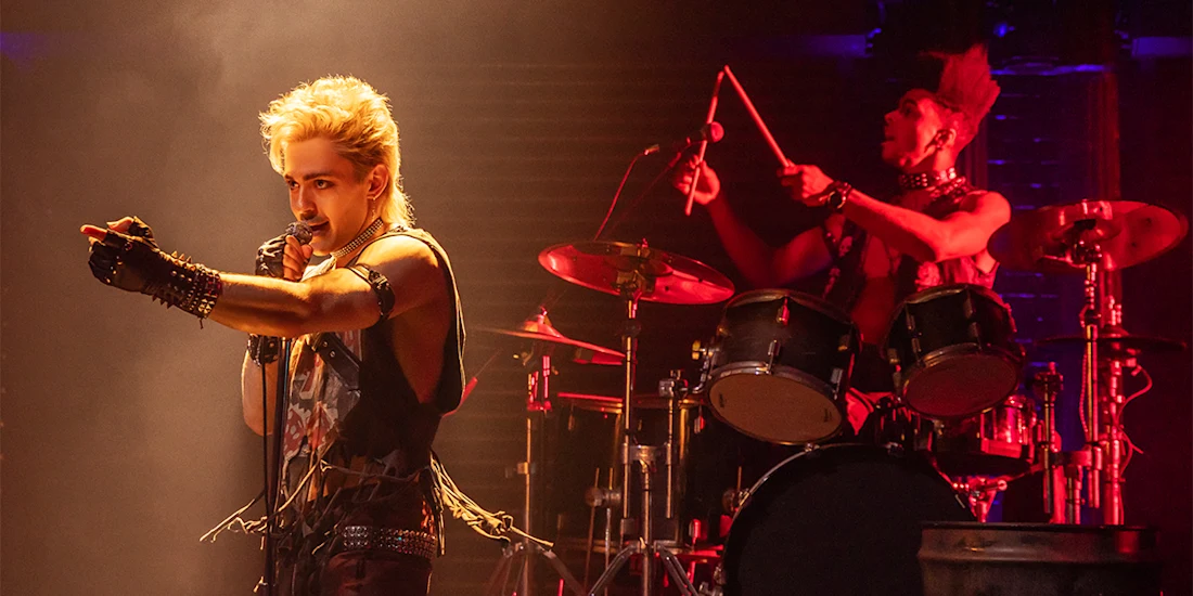 Two musicians perform on stage; the singer points forward while holding a microphone, and the drummer plays behind a drum set under dramatic lighting.