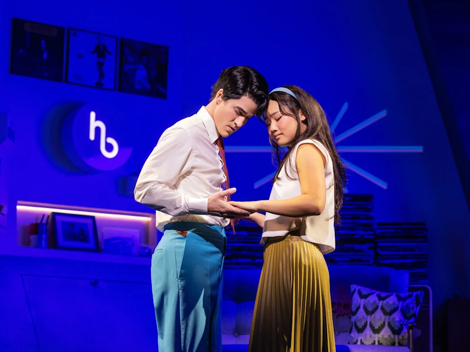 Two people stand close together on stage, holding hands and looking down, with blue lighting and modern decor in the background.