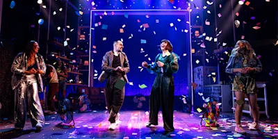 Four people stand and dance on a stage under colorful confetti and dramatic lighting, with props and shelves in the background, suggesting a lively performance scene.